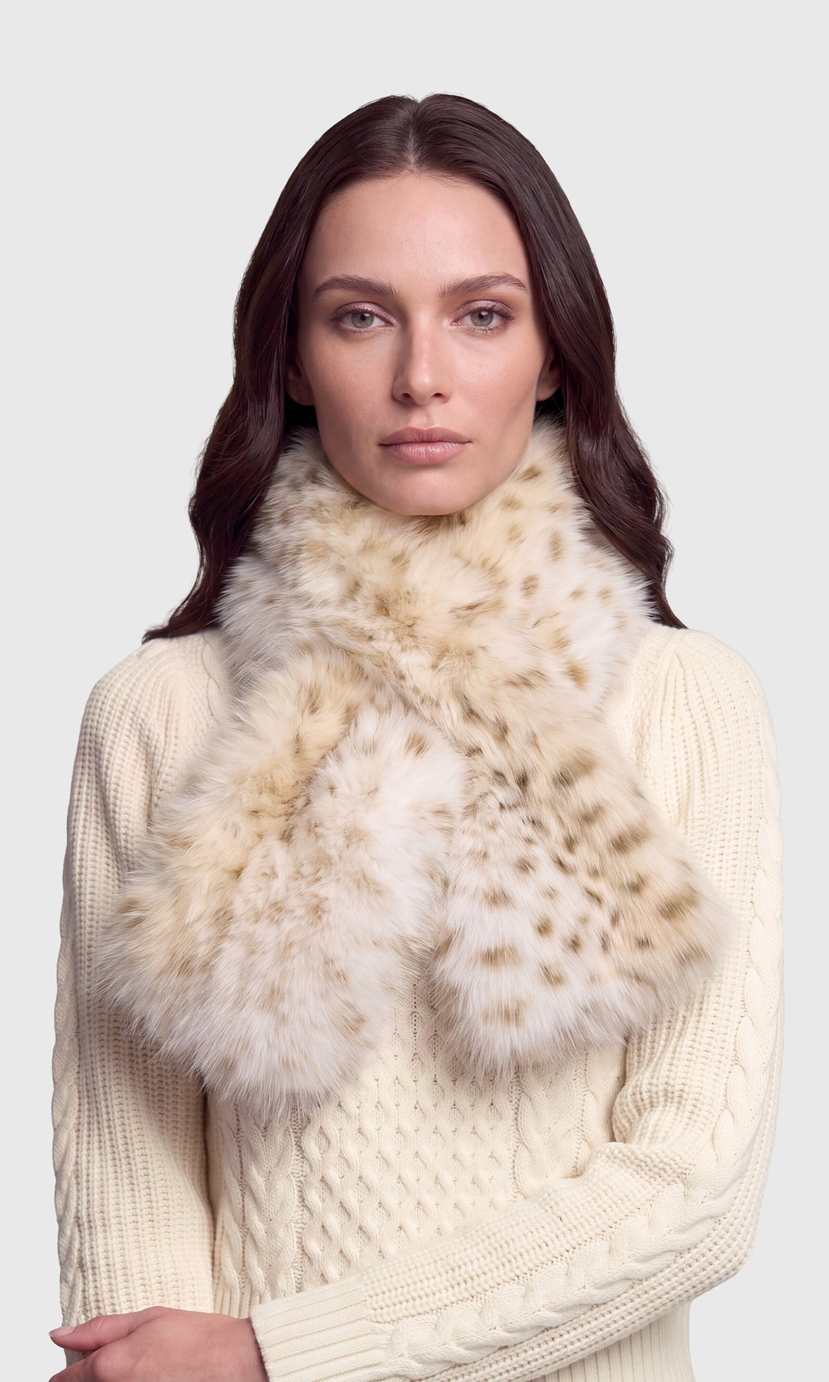 Model wearing a cream cable-knit sweater and a thick beige and brown spotted faux-fur scarf.