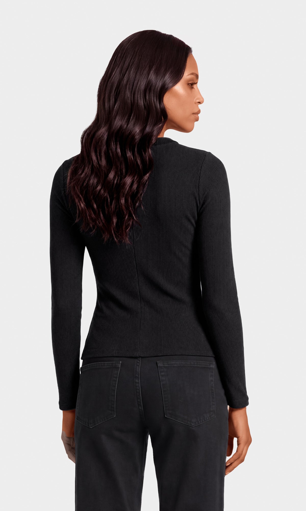 A woman shown from the back wearing a fitted black long-sleeve top and dark jeans against a light gray studio background.
