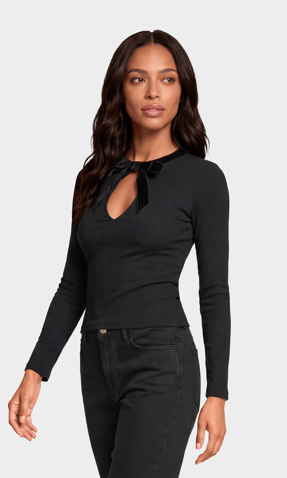 Side view of model wearing a black long-sleeve knit top with a keyhole cutout and a velvet bow detail at the collar, paired with black jeans.