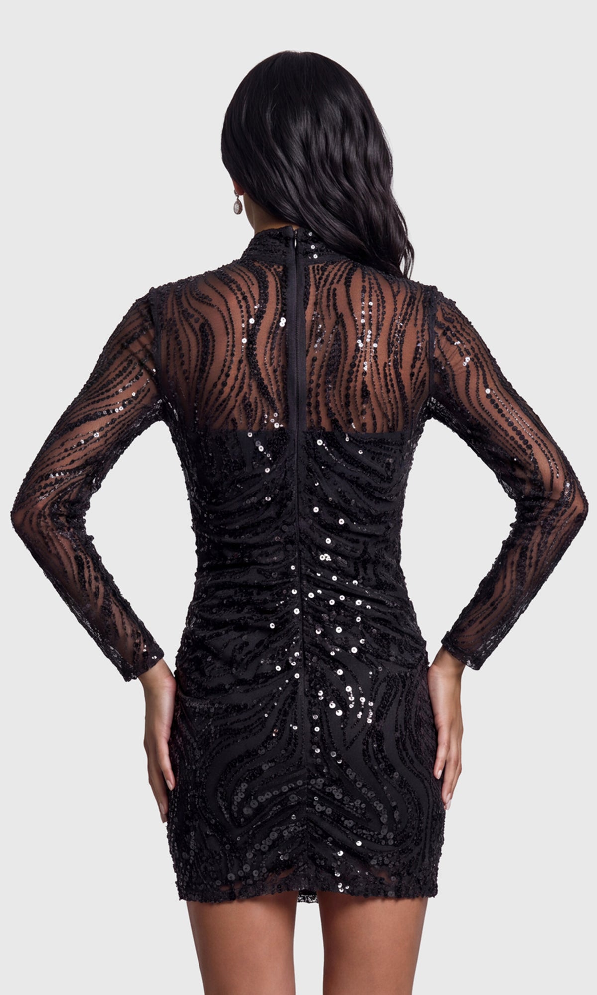 Back view of model wearing a black long-sleeve mini dress with sheer lace sleeves, a high mock neck, and all-over sequined detailing.