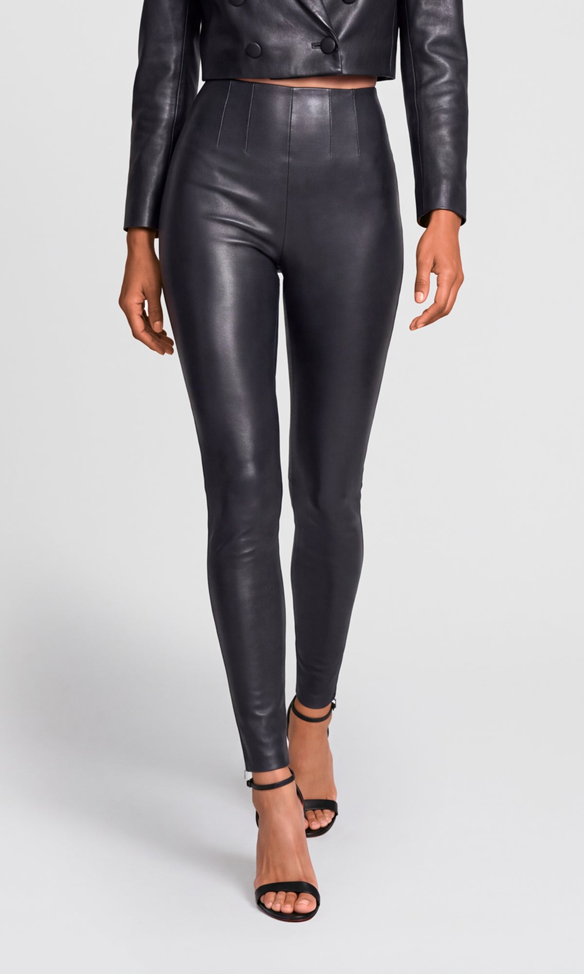 A model wearing high-waisted, black faux-leather leggings and a matching cropped double-breasted jacket, paired with black strappy high-heeled sandals.