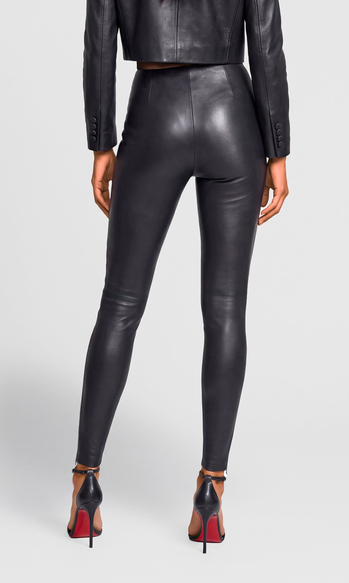Rear view of a model wearing black faux-leather leggings, a black cropped jacket, and black high heels with red soles.