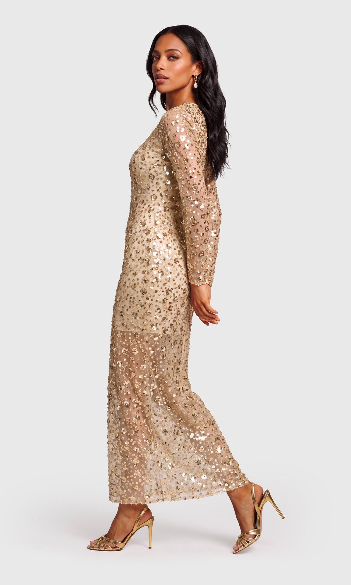 Side view of model wearing a long-sleeve, floor-length champagne gold sequined gown with a thigh-high slit.
