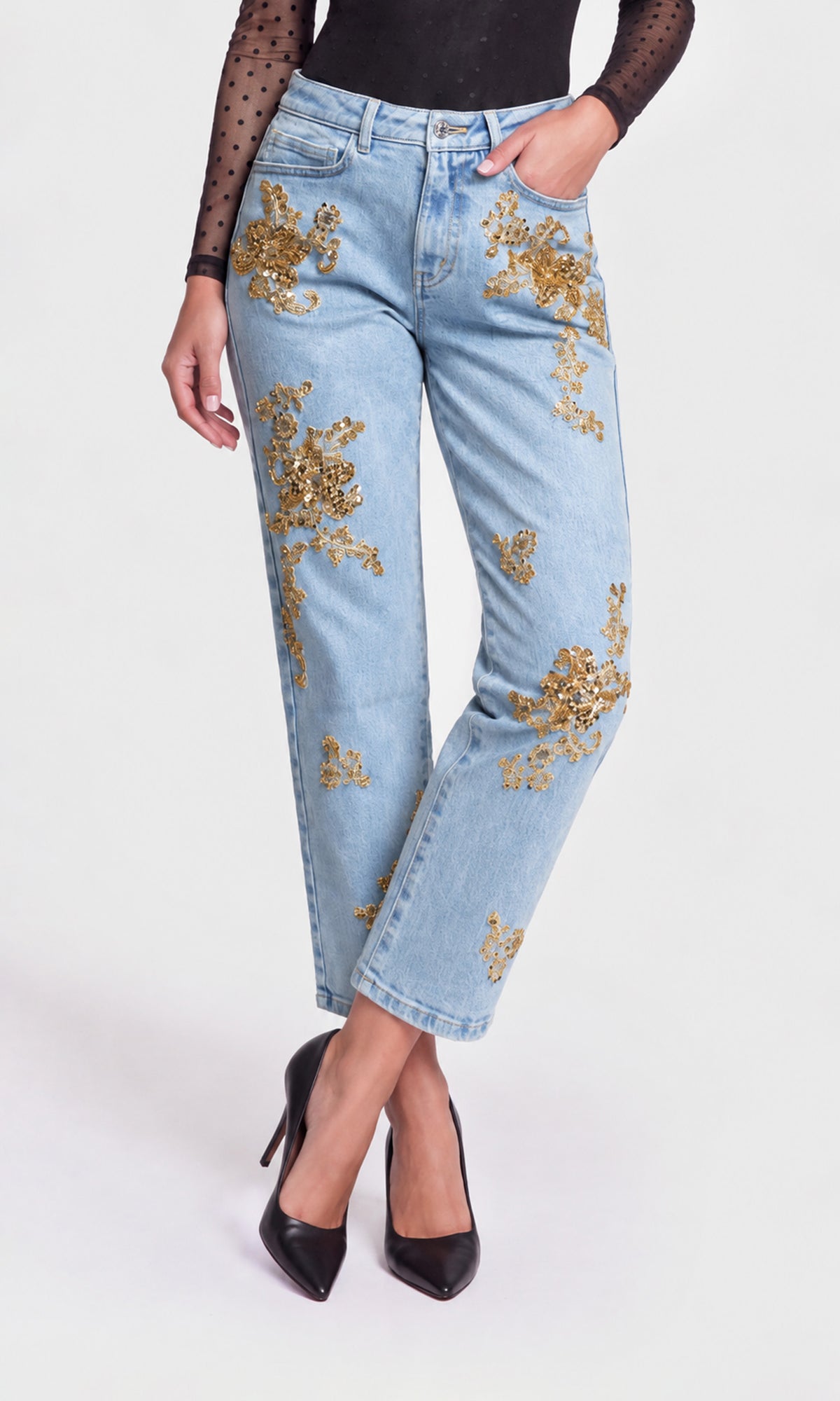 A model wearing light-wash blue jeans with gold sequined floral embroidery, styled with a black sheer long-sleeve top.
