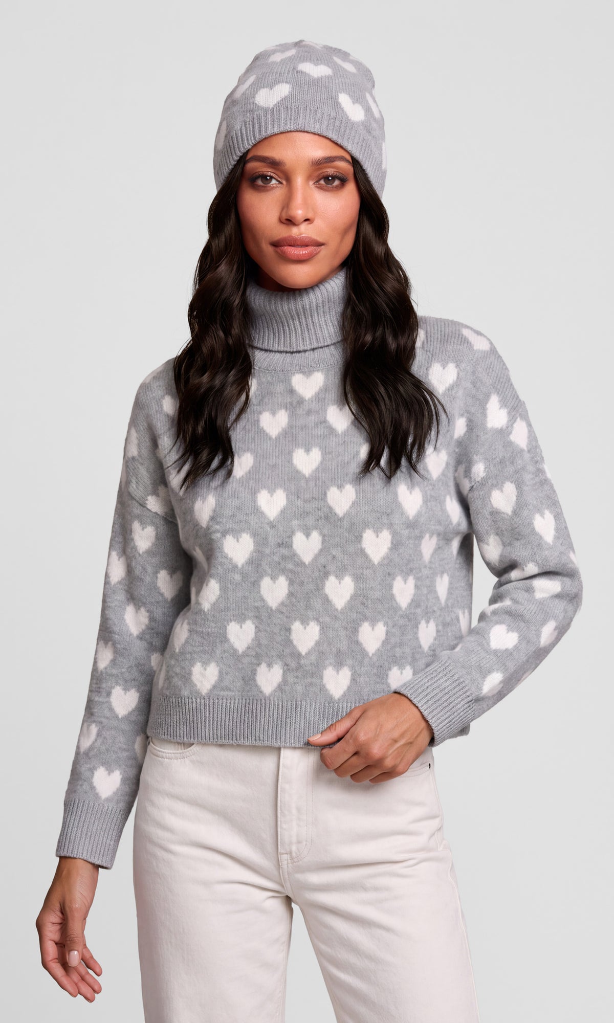 A woman wearing a light gray knit turtleneck sweater with a white heart pattern, a matching gray beanie, and off-white pants.