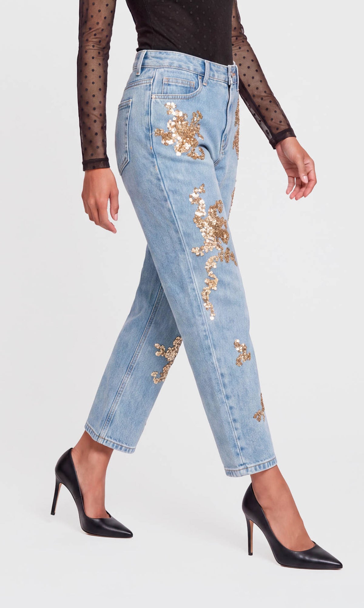 Side view of model wearing light-wash blue jeans with gold sequined floral embroidery, styled with a black sheer long-sleeve top.