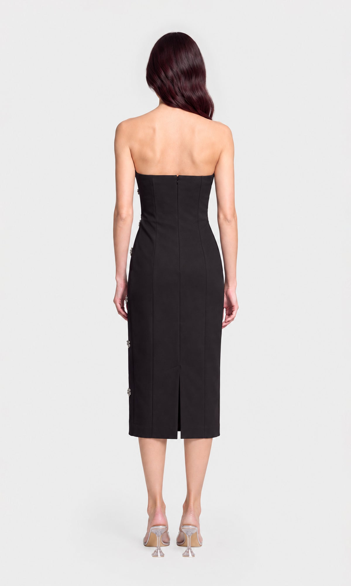 A woman viewed from the back wearing a strapless black midi dress with a fitted waist and a small back hem slit, paired with clear high-heeled sandals.