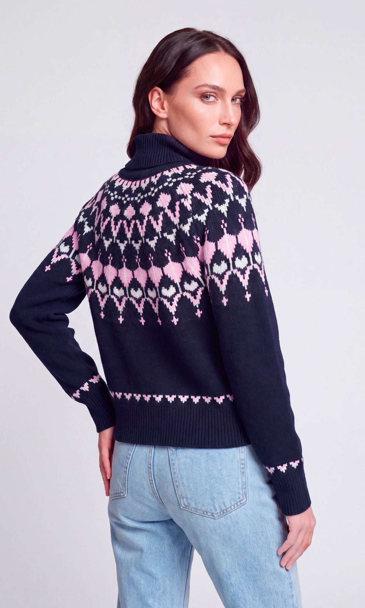 Back view of model wearing a navy blue turtleneck sweater with a pink and white geometric pattern on the back and sleeves, paired with light blue jeans.