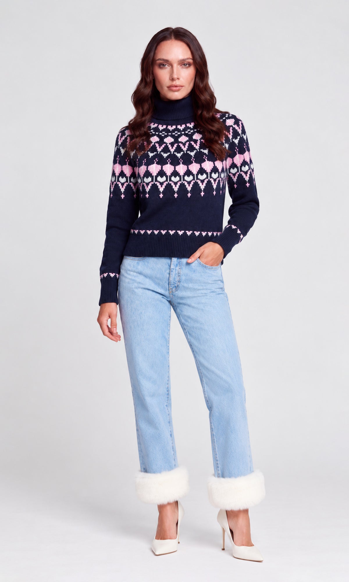 A woman modeling a navy blue knit sweater with a pink and white geometric pattern, paired with light blue jeans featuring white faux-fur trim at the cuffs and white pointed-toe heels.