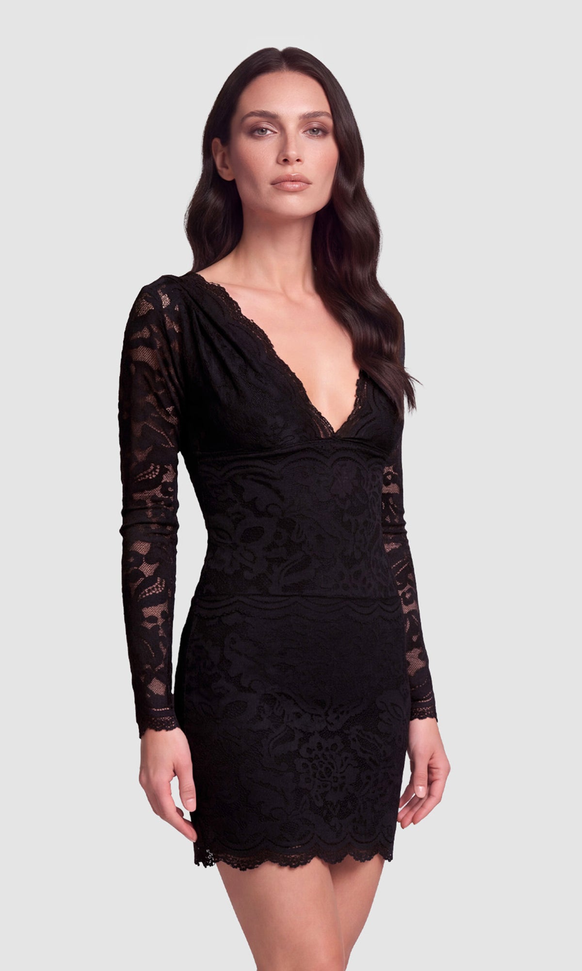 A woman modeling a black long-sleeve lace mini dress with a deep V-neckline, photographed against a solid light gray studio background.