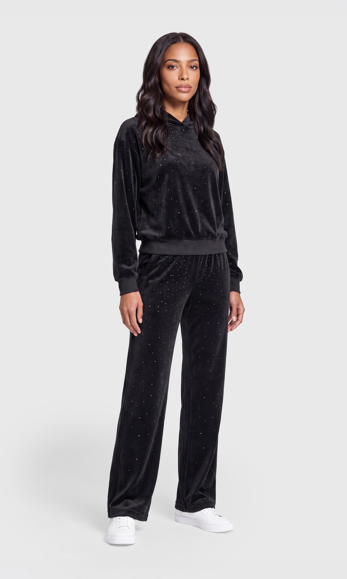 A woman modeling a matching black velour tracksuit with a long-sleeve top and wide-leg pants, styled with white sneakers.