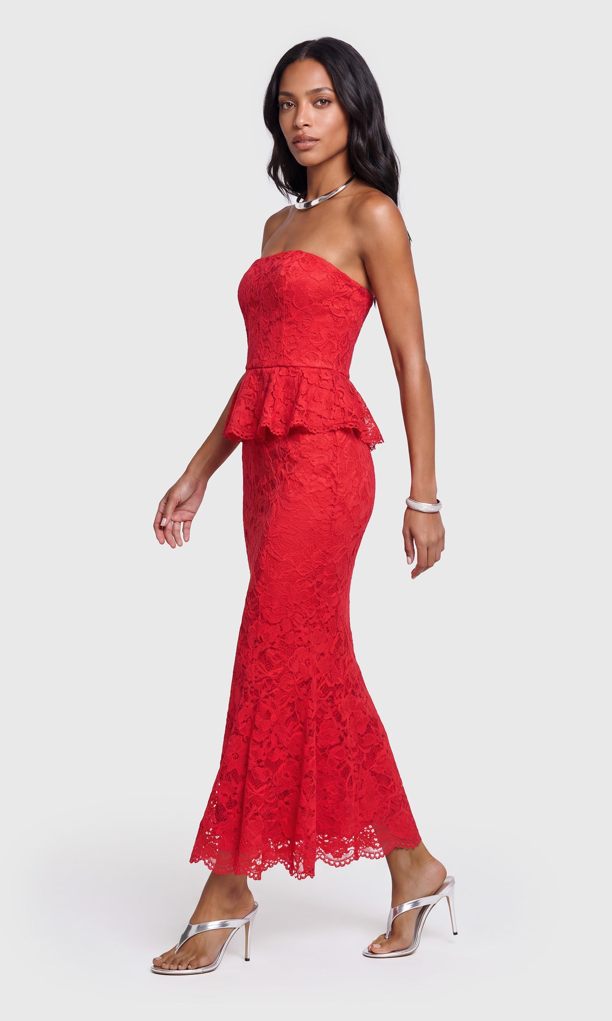 Side view of model wearing a strapless red lace maxi dress with a peplum waist and flared lace hem, paired with clear high-heel sandals.