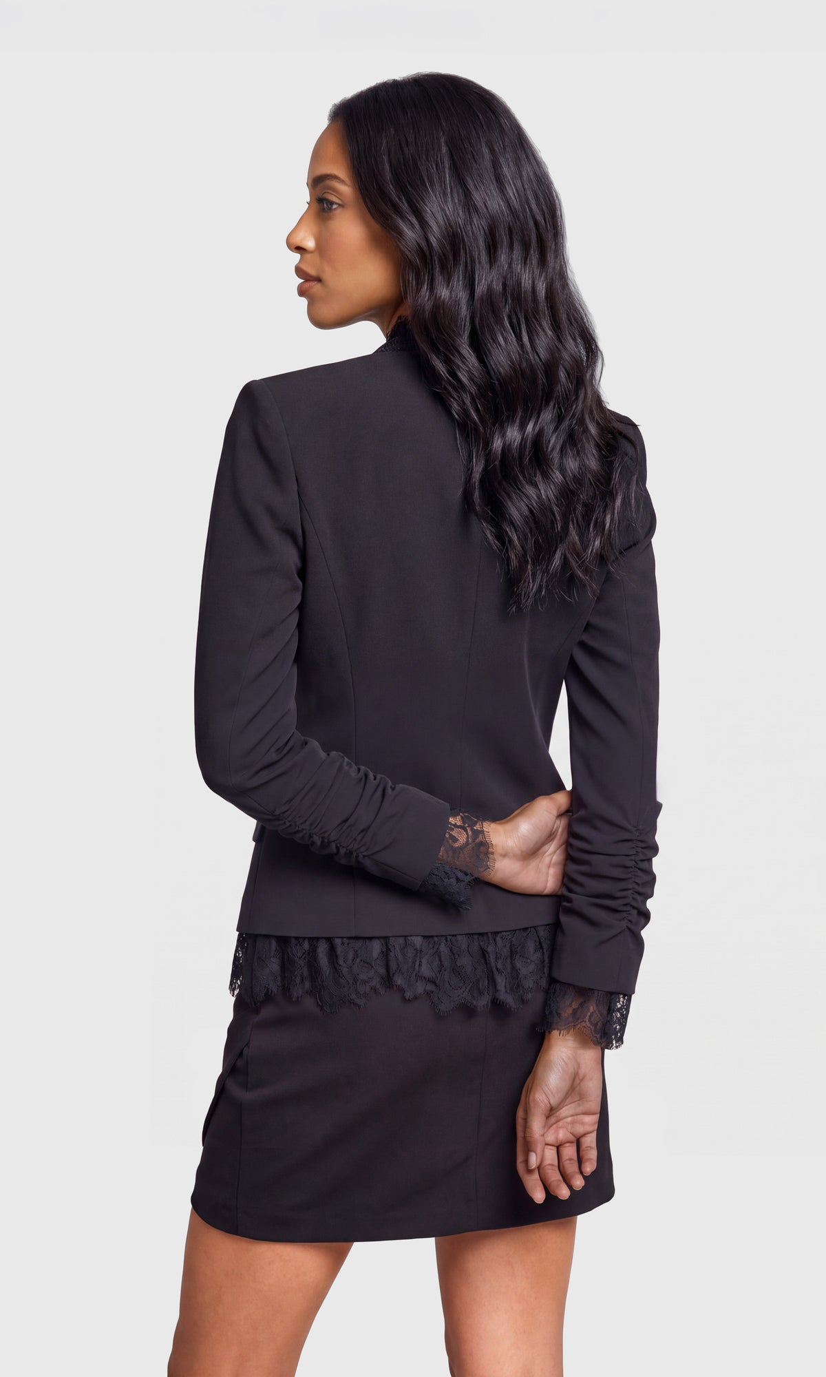 Back view of model wearing a black long-sleeve blazer with lace trim on the cuffs and hem, paired with a matching black mini skirt.