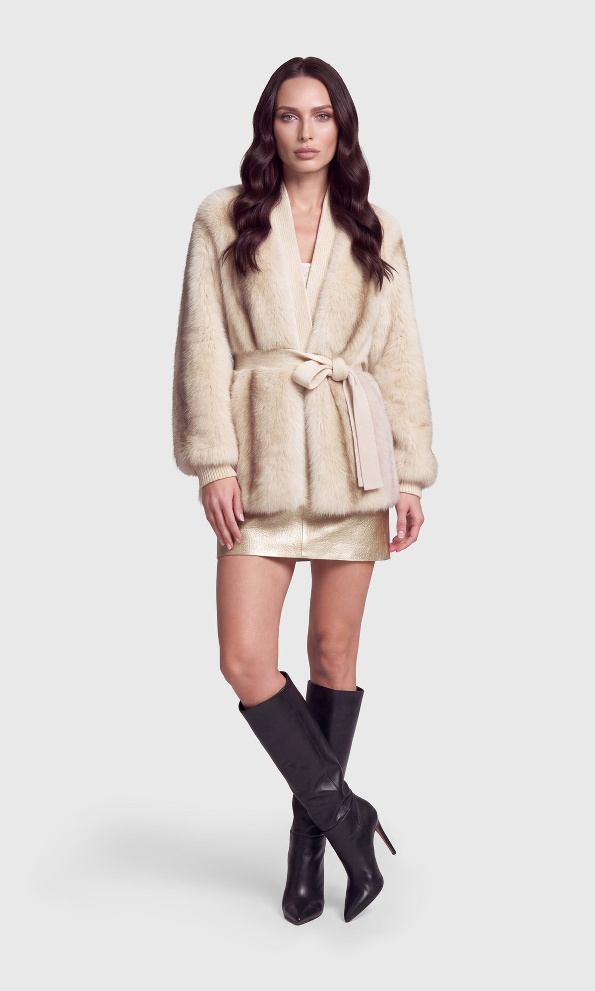 A woman modeling a cream-colored faux fur wrap coat with a tie belt, paired with a metallic gold mini skirt and black knee-high leather boots.