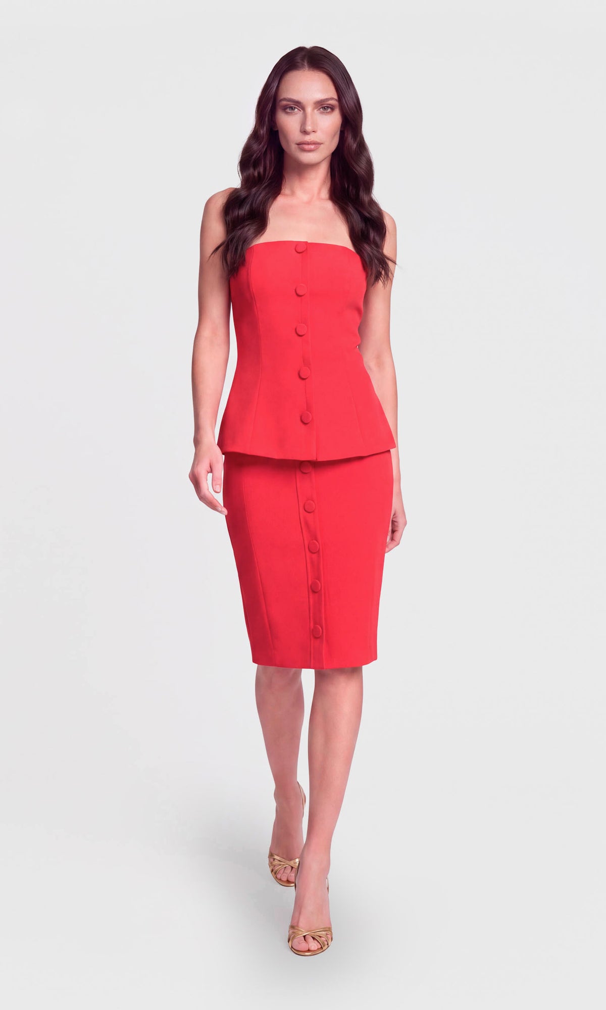 A woman modeling a bright red two-piece set consisting of a strapless peplum-style top with front buttons and a matching knee-length pencil skirt with front buttons.