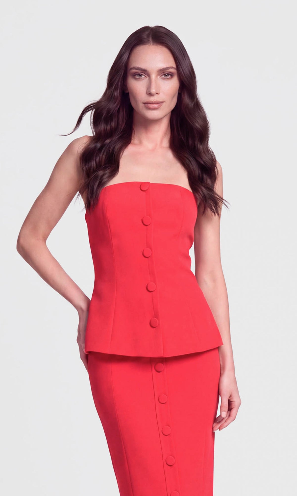 Model wearing a red strapless peplum top with a matching skirt with fabric covered buttons down the front.