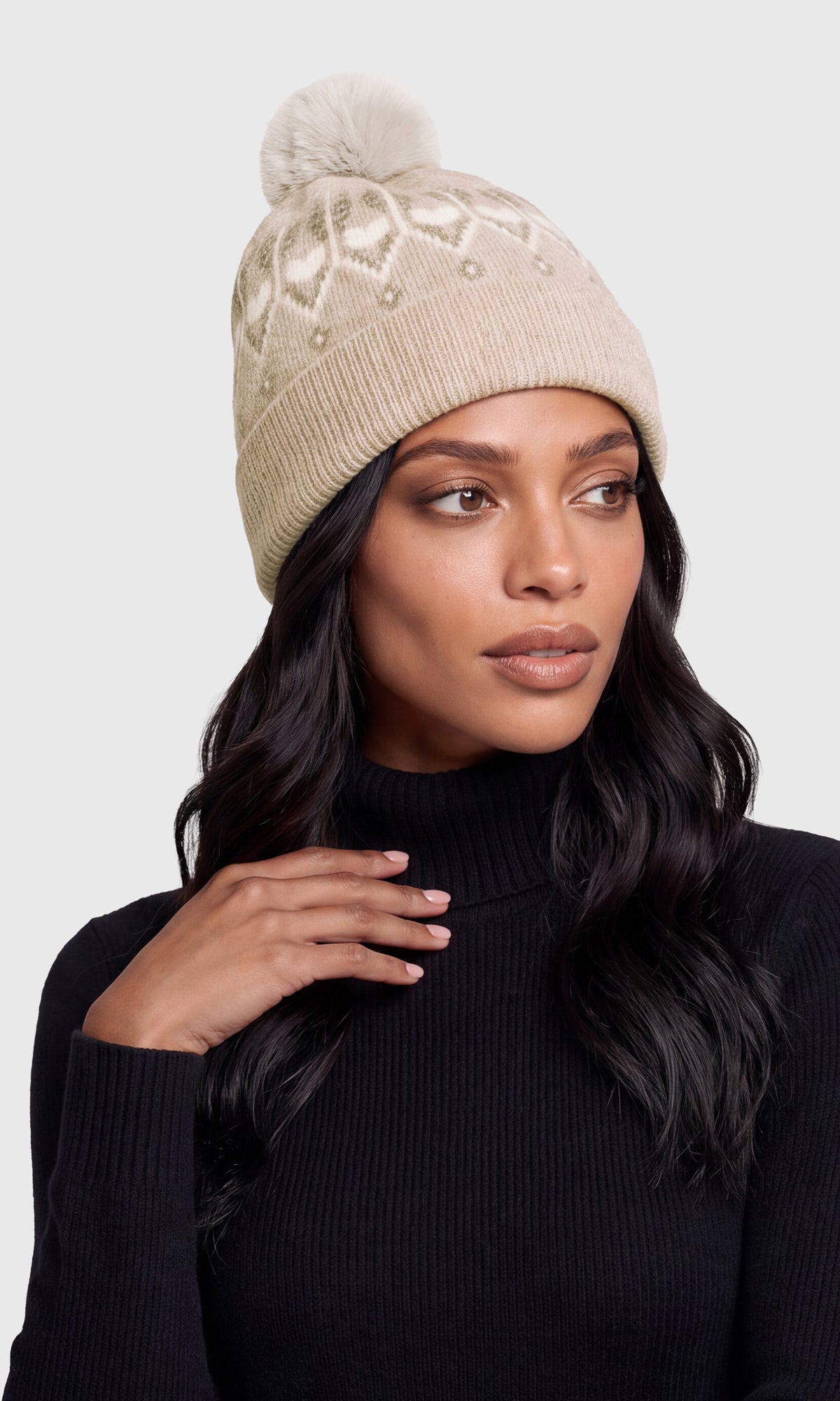 A woman wearing a beige knit beanie with a pom-pom and subtle geometric knit pattern, paired with a black ribbed turtleneck sweater.