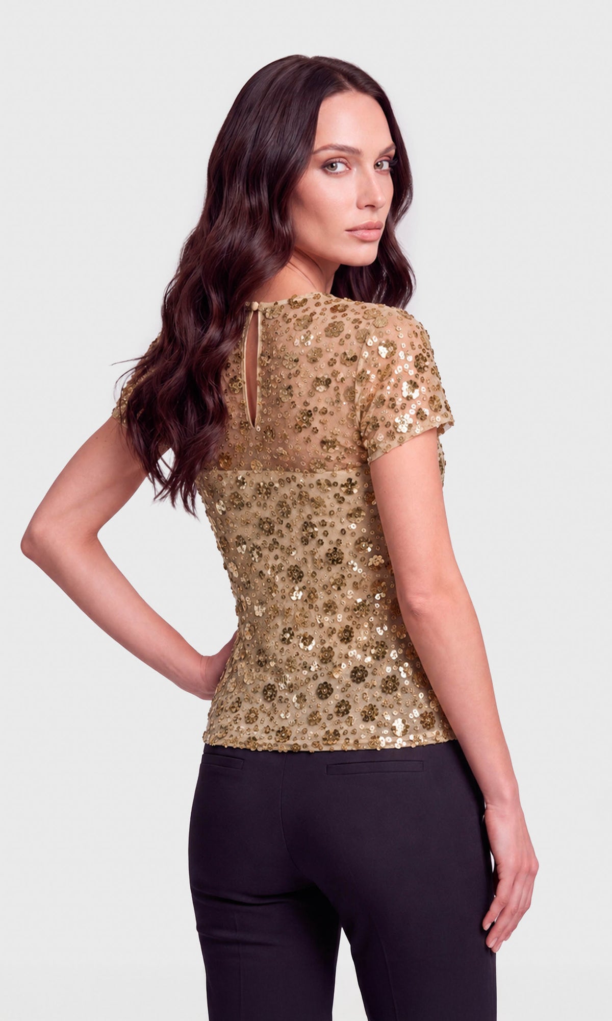 A woman shown from the back wearing a short-sleeve gold sequined top and black pants against a light gray studio background.