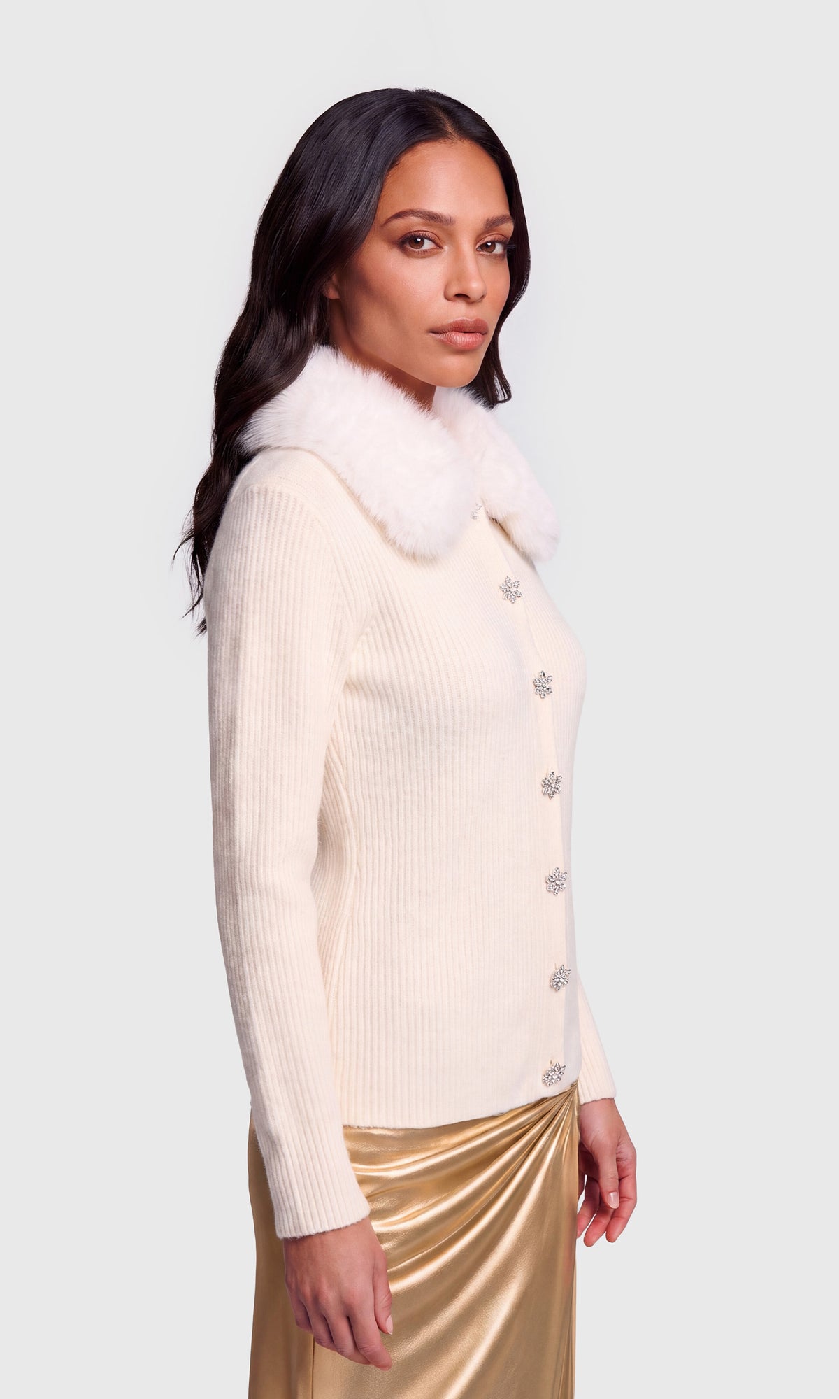 Side view of model wearing a cream ribbed knit cardigan with a fluffy white faux-fur collar and decorative crystal-like buttons, paired with a metallic gold skirt.
