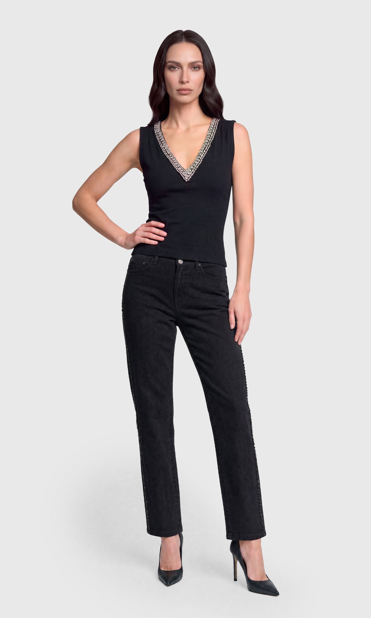 A woman modeling a black sleeveless V-neck top with a silver beaded trim and black pants against a light gray studio background.