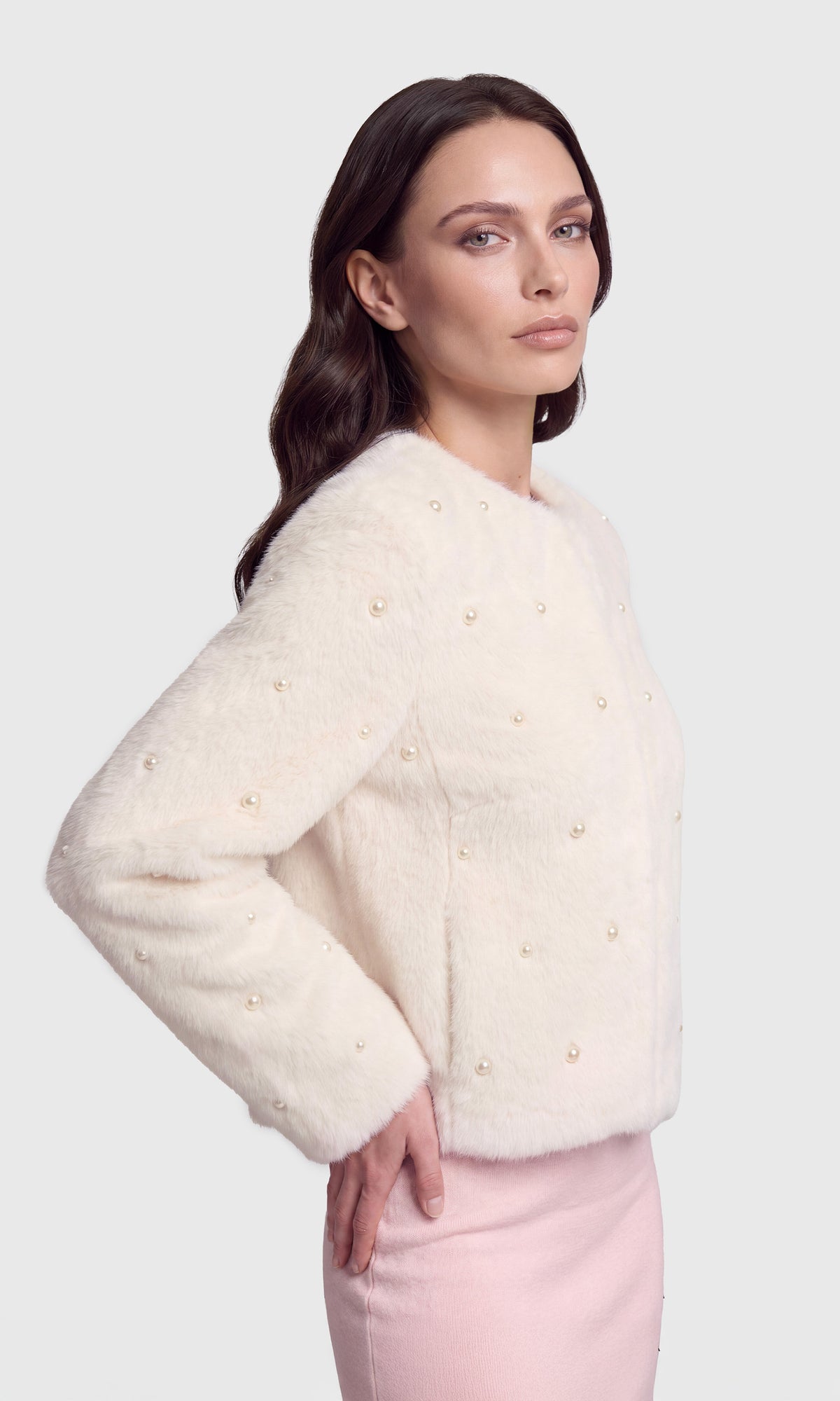 A woman modeling a cream faux fur cropped jacket with pearl embellishments, paired with a light pink dress.