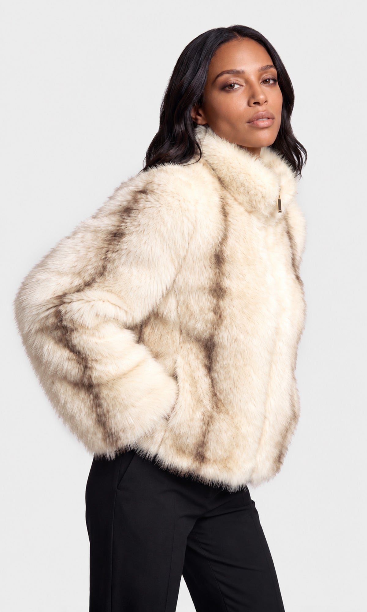 Side view of model wearing a cream-colored faux fur jacket with a high collar and a subtle brown mottled pattern, paired with black pants.
