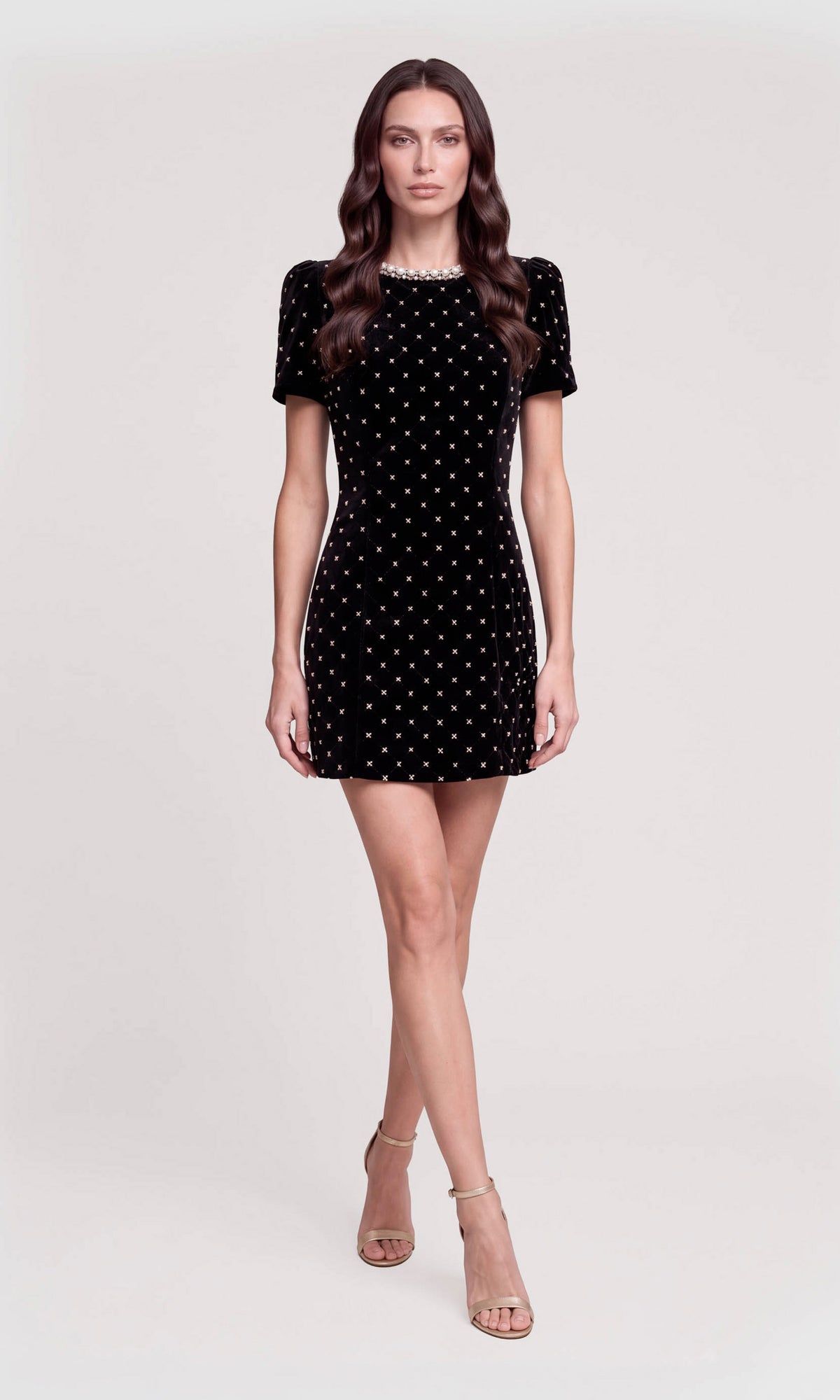 A woman modeling a short-sleeve black velvet mini dress with a white polka dot pattern and a beaded neckline, paired with nude high-heeled sandals.