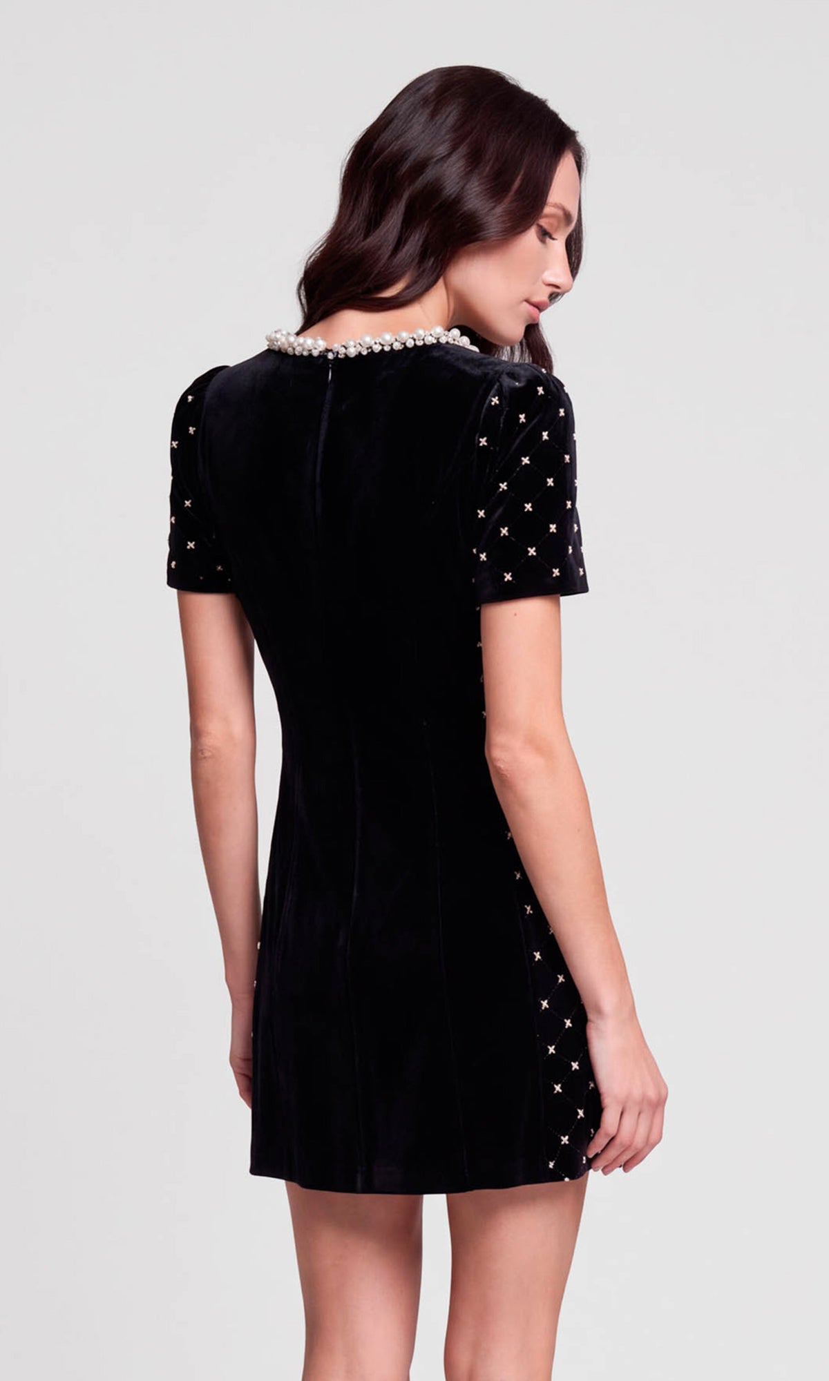 Back view of model wearing a short-sleeve black velvet dress from the back, featuring a pearl-trimmed neckline and silver rhinestone embellishments on the sleeves.