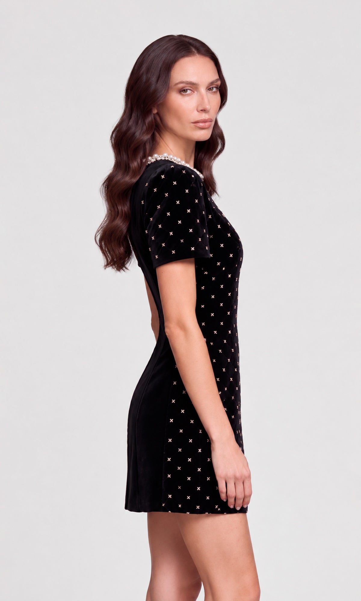 A woman modeling a short black velvet dress with gold polka dots and a beaded neckline, shown from a side profile against.