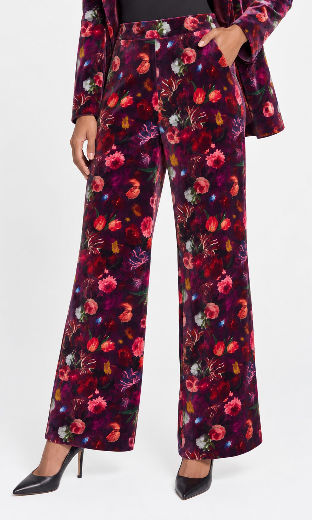 A model wearing a pair of wide-leg trousers and a matching long-sleeve top in a dark purple velvet fabric with a vibrant multicolor floral print.