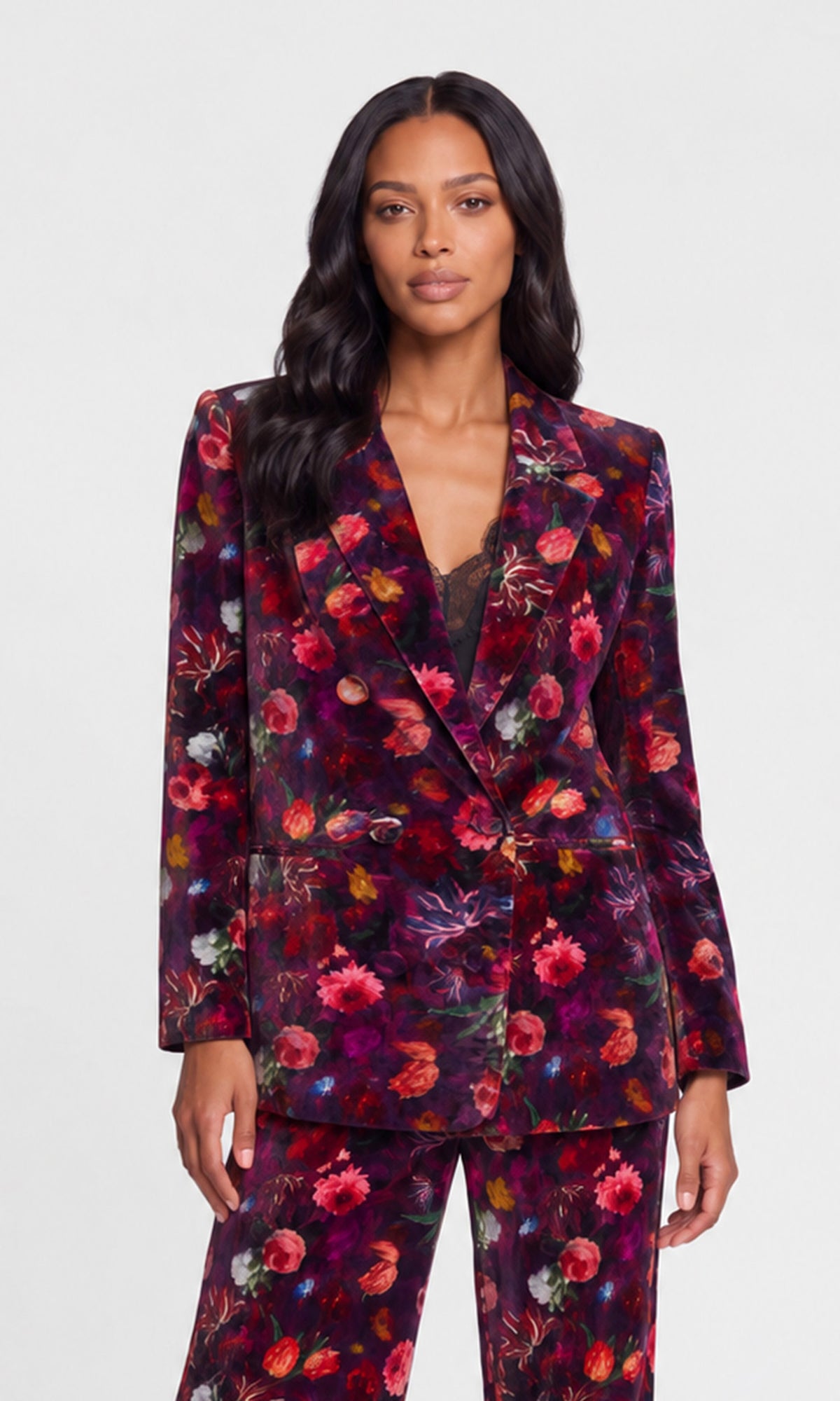 A woman modeling a dark floral velvet blazer and matching trousers against a light gray studio background.