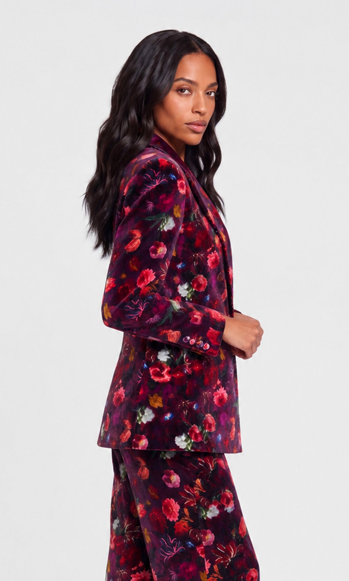 Side view of model wearing a dark floral velvet blazer and matching trousers against a light gray studio background.