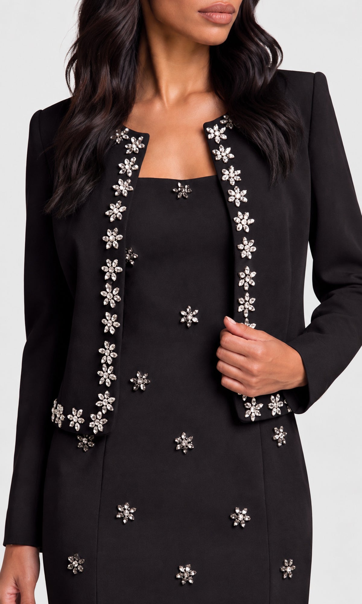 A woman wearing a black tailored jacket and matching dress, both decorated with silver-toned floral embellishments.