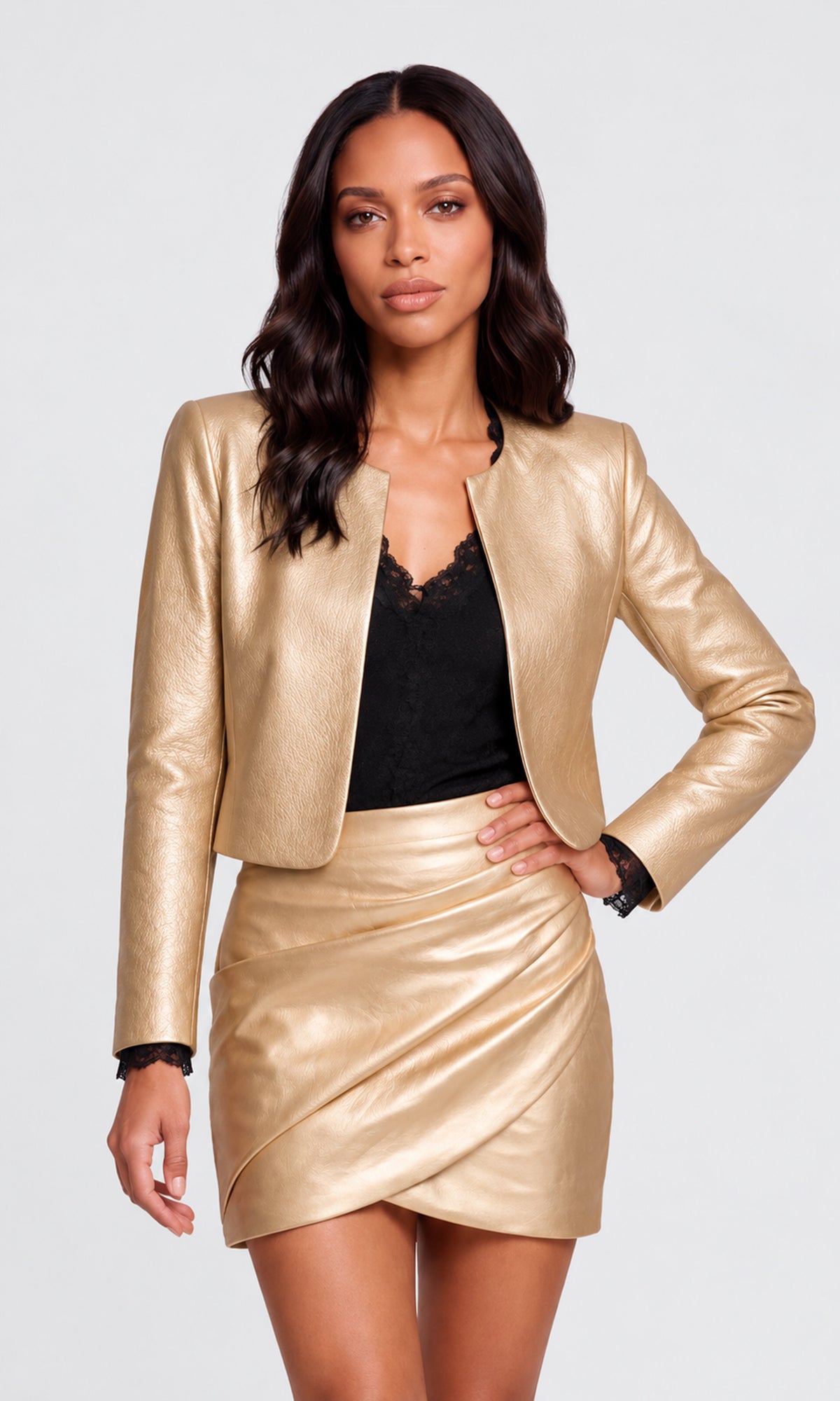 A woman modeling a metallic gold faux-leather jacket and matching mini skirt set, worn over a black lace-trim top, against a light gray studio background.