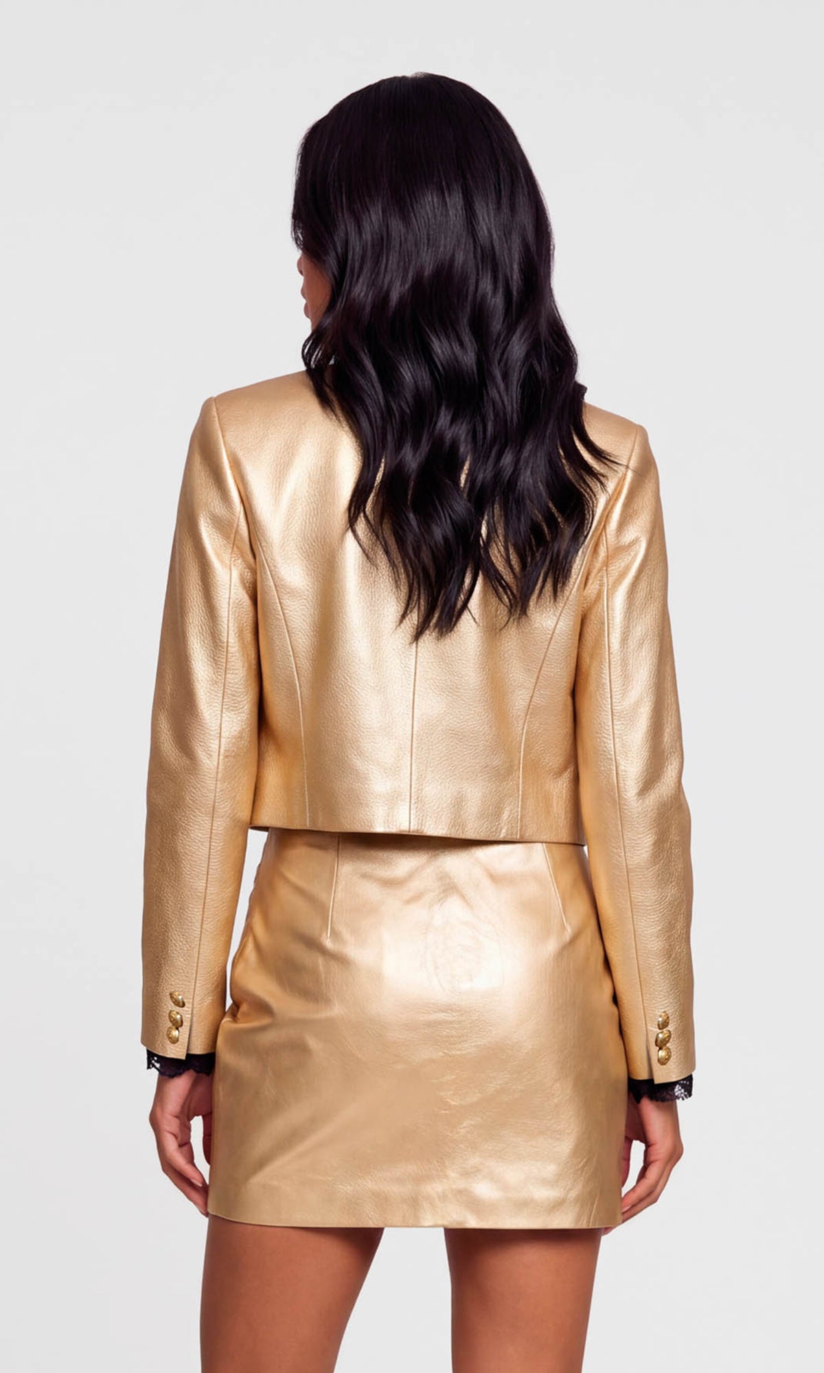 A woman shown from the back wearing a metallic gold faux-leather cropped blazer and matching mini skirt against a light gray studio background.