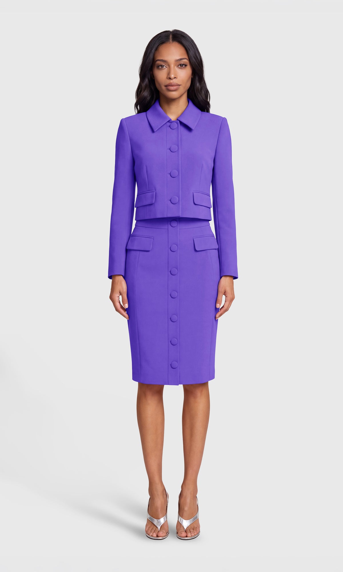 Model wearing a bright purple two-piece suit set consisting of a long-sleeve cropped jacket with a pointed collar and matching button-front pencil skirt.