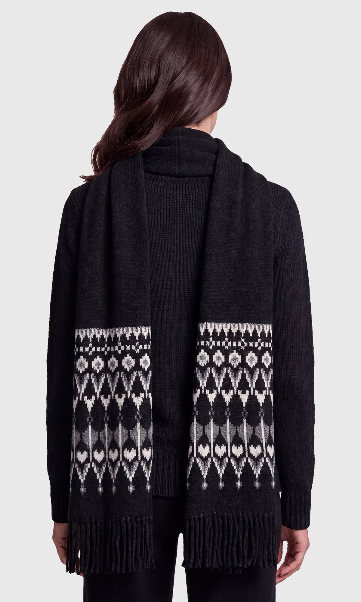 A woman shown from the back wearing a black knit scarf with a white geometric Fair Isle pattern and fringe, over a black ribbed sweater.