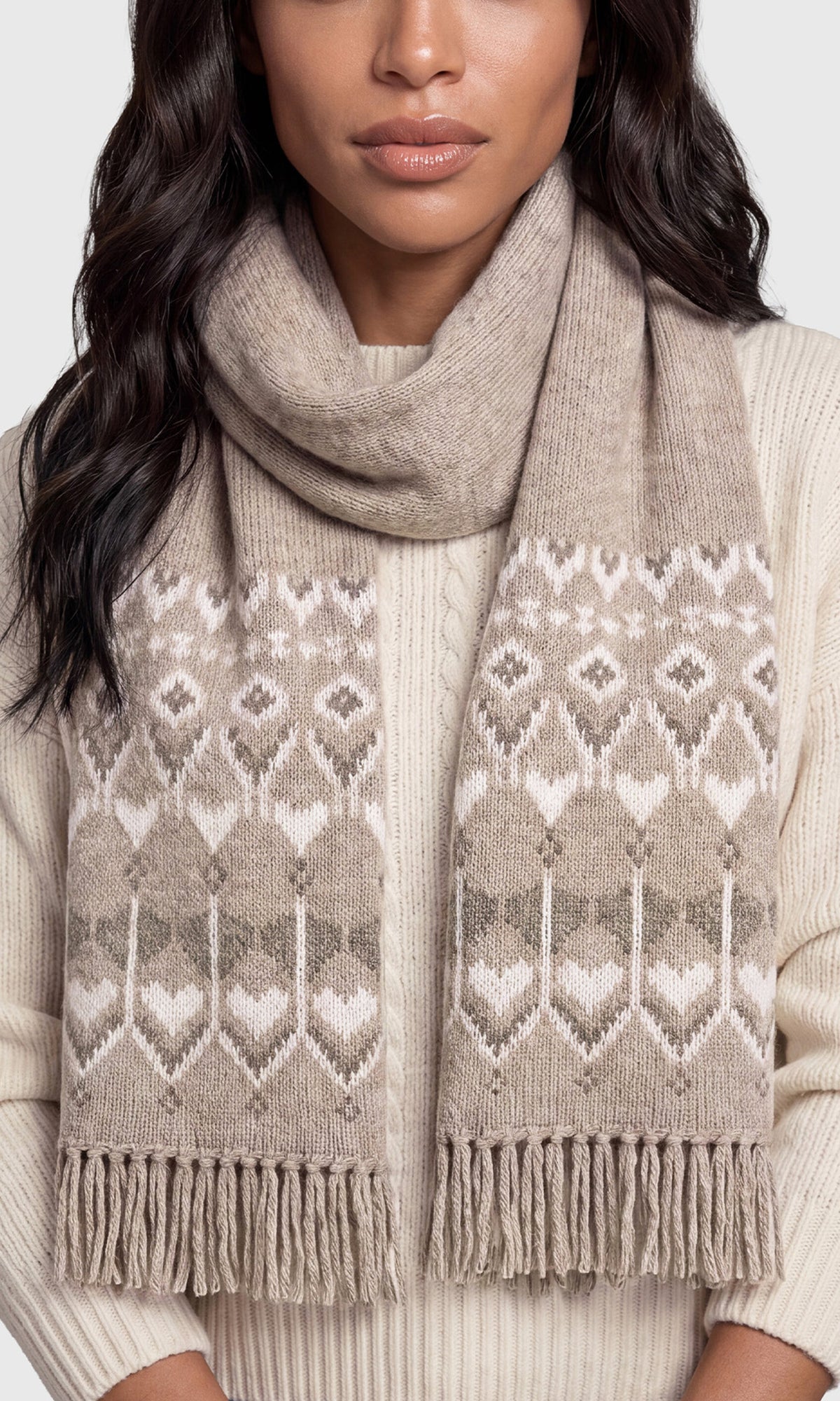 A close-up of a woman wearing a chunky knit scarf in taupe and cream with a geometric Fair Isle pattern and fringed ends, paired with a cream ribbed sweater.
