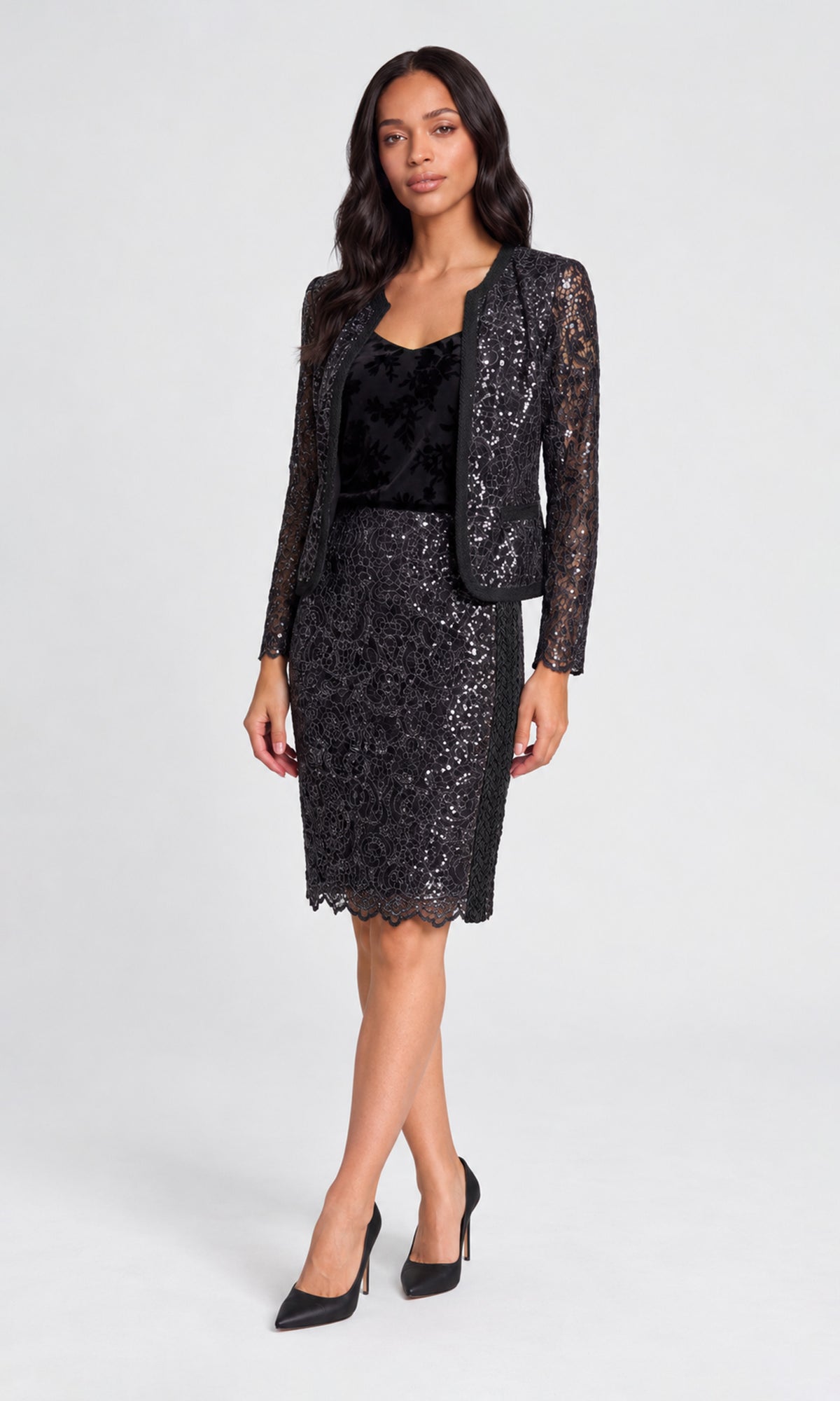 A woman models a black sequined two-piece set consisting of a long-sleeve blazer and a matching knee-length skirt, paired with a black velvet top and black pointed-toe heels.