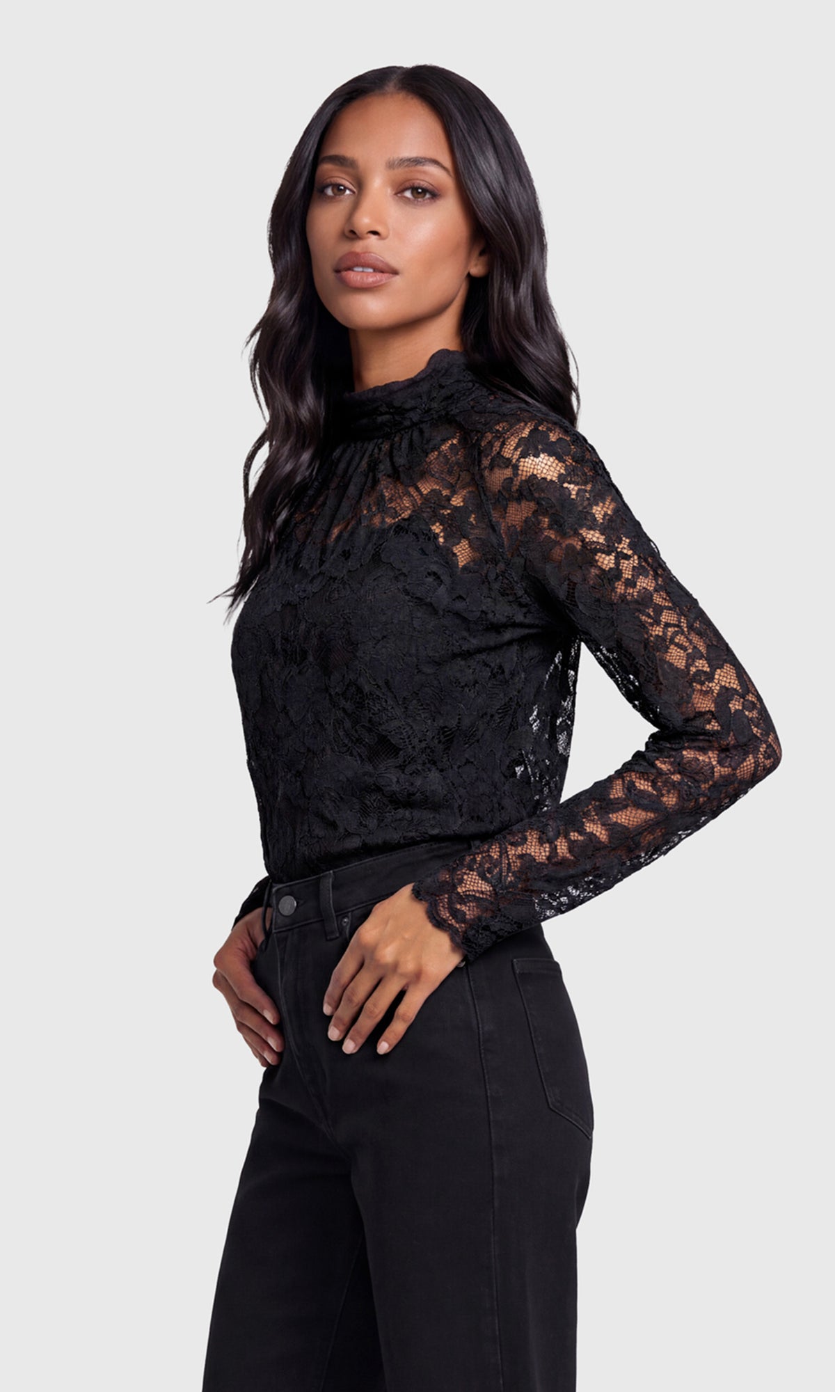 Side view of model wearing a black lace, mock-neck top with black jeans.