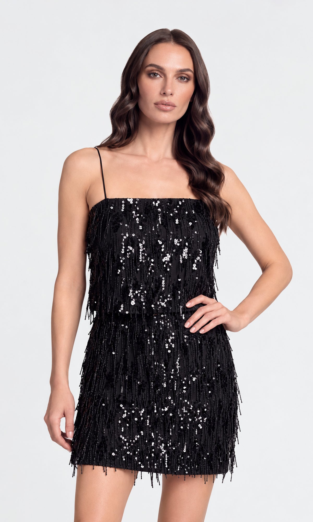 A woman modeling a black sequined mini skirt and matching top with fringe detailing and thin spaghetti straps against a light gray studio background.