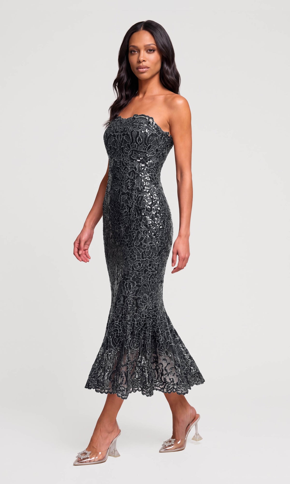 Side view of model wearing a strapless black lace and sequin mermaid gown with clear high-heeled sandals.