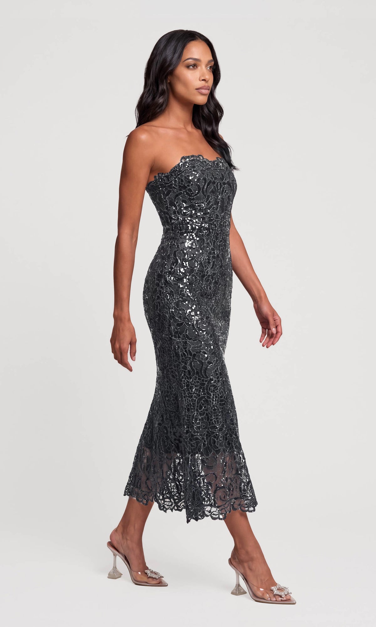 Side view of model wearing a strapless black lace and sequin mermaid gown with clear high-heeled sandals.