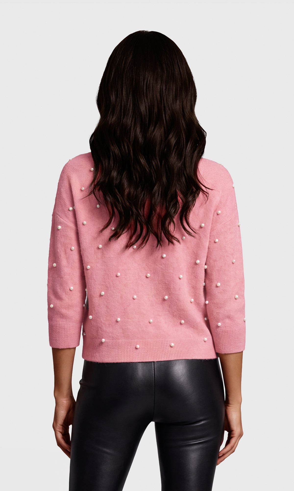 Back view of model wearing a pink knit sweater with small pearl-like embellishments and black faux-leather pants.
