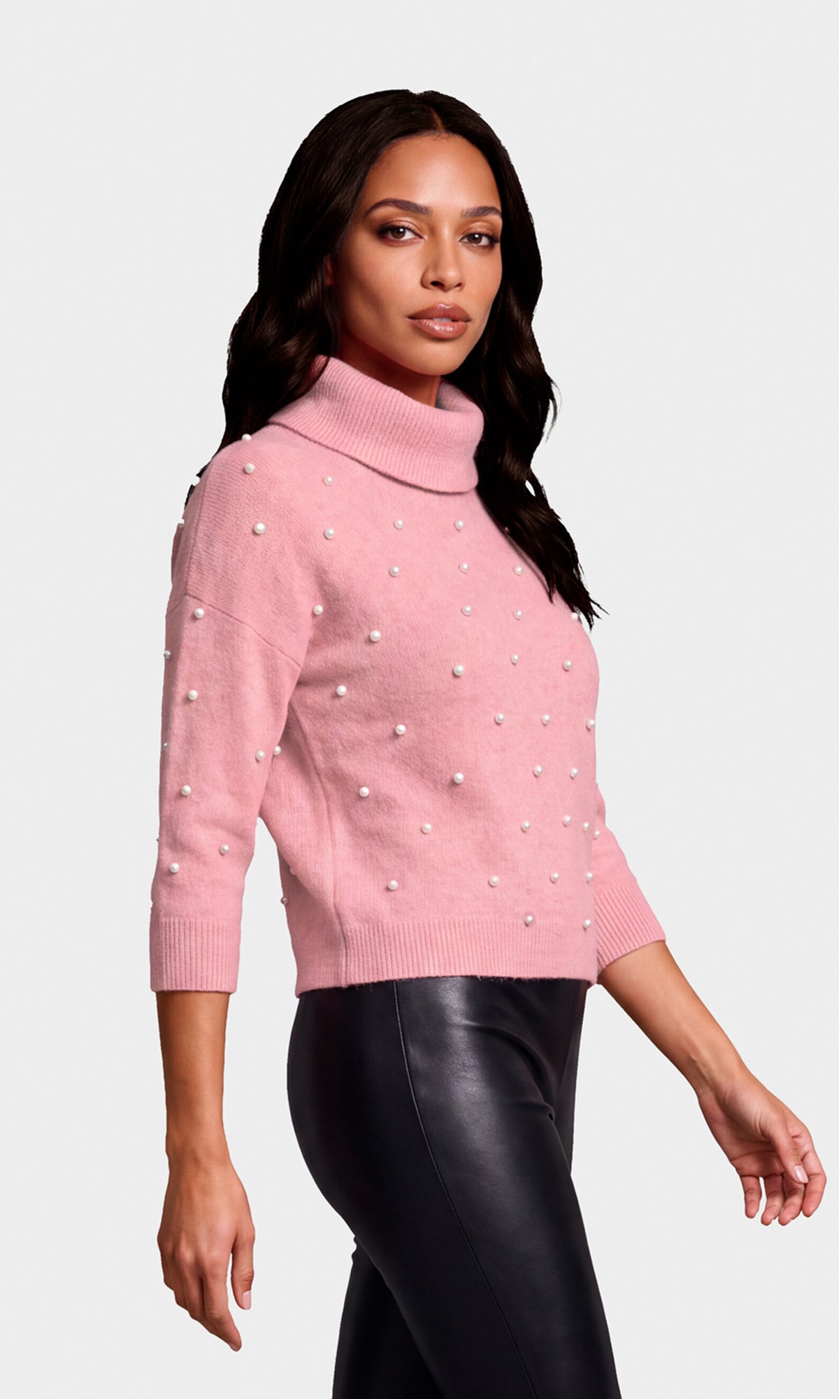 Side view of model wearing a light pink turtleneck sweater adorned with small white pearl embellishments, paired with black faux-leather pants.