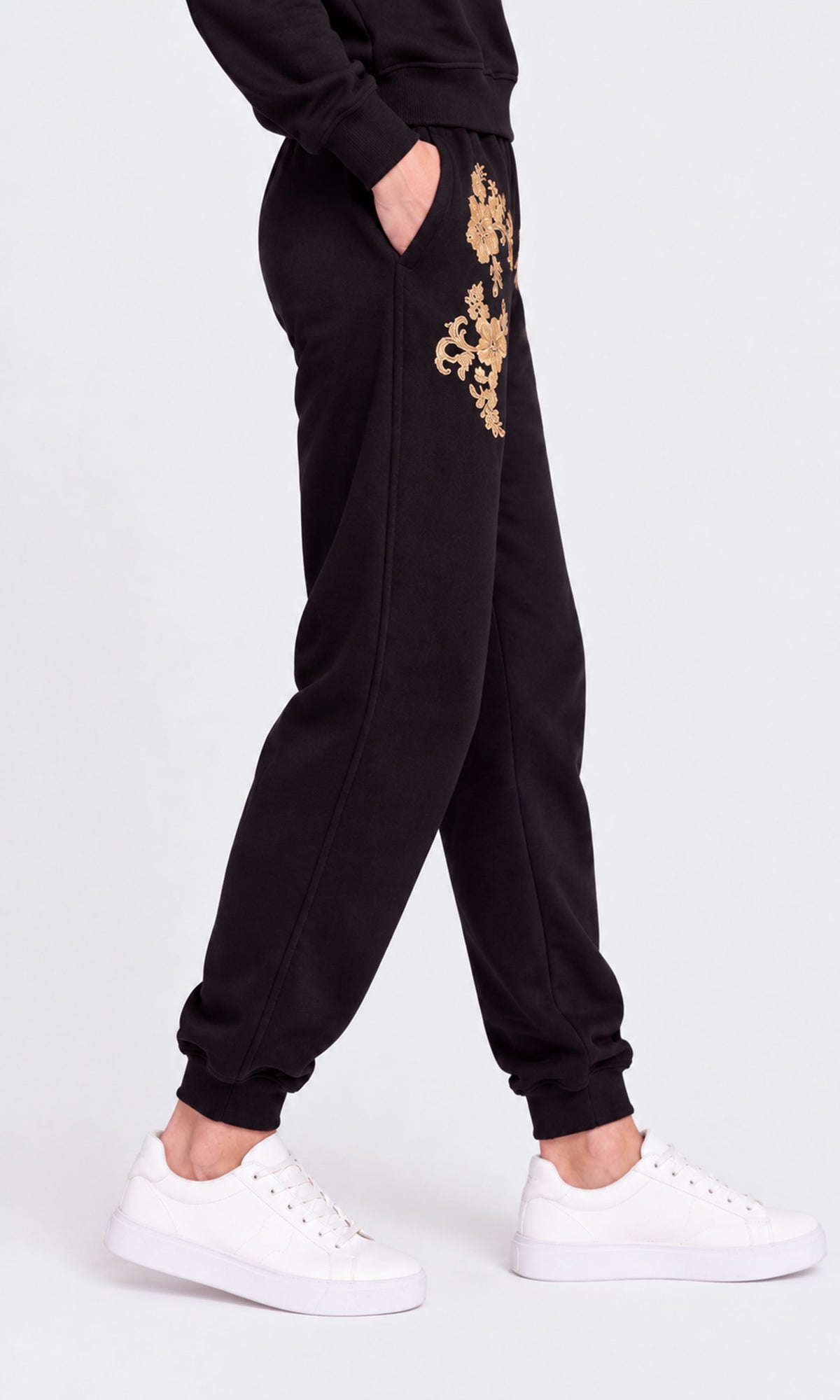Side view of model wearing black sweatpants with gold floral embroidery on the upper thighs, paired with white sneakers.