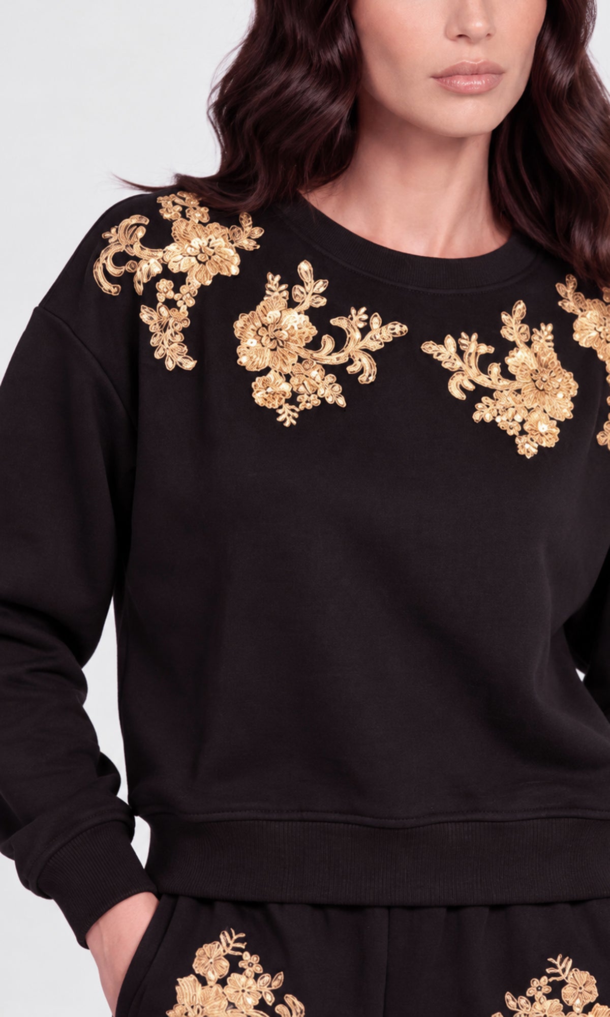A close-up of a model wearing a black crewneck sweatshirt with ornate gold floral embroidery across the shoulders and upper chest.