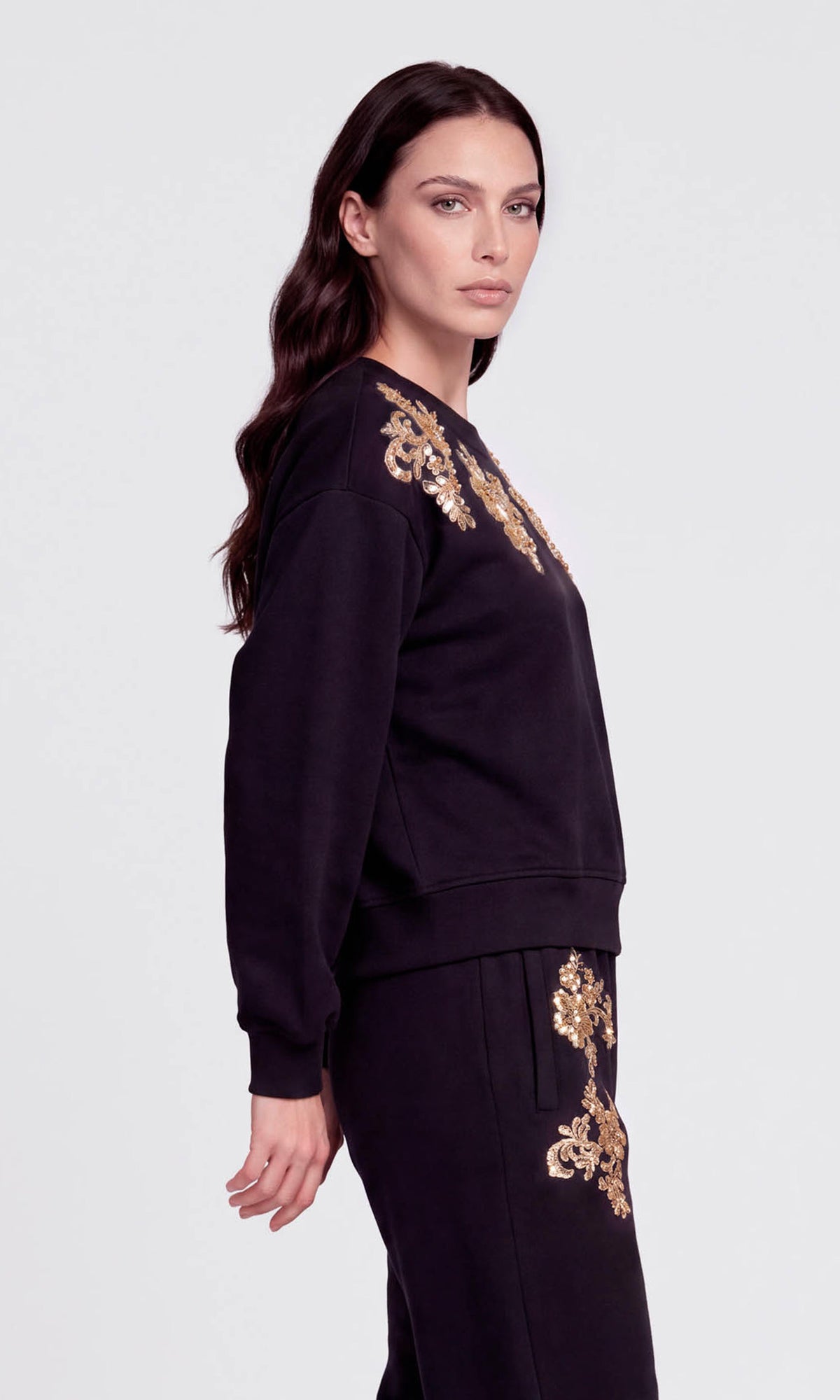 Side view of model wearing a black crewneck sweatshirt and matching sweatpants set, both decorated with ornate gold floral embroidery on the shoulders and upper thighs.