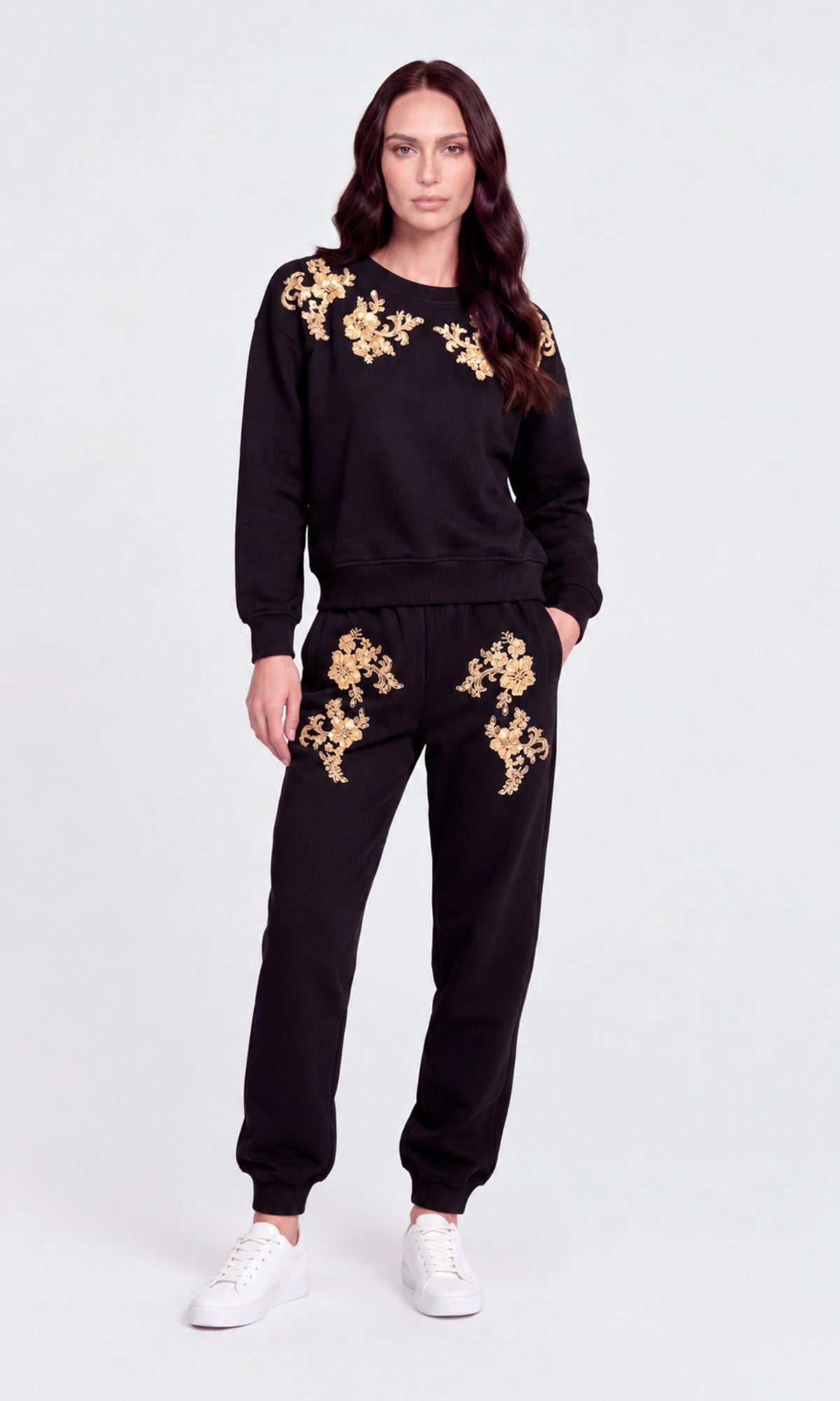 A woman modeling a black sweatshirt and jogger pants set with gold floral embroidery on the chest and thighs, paired with white sneakers.