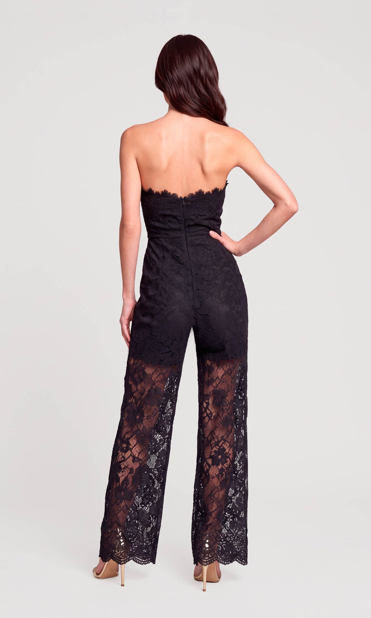 A woman shown from the back wearing a strapless black lace jumpsuit with wide legs and nude heels.